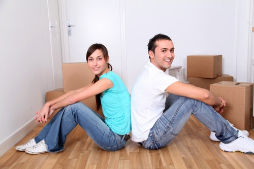 A young woman and a young man are sitting back-to-back on a light wooden floor inside a clean, white-walled room, surrounded by several cardboard moving boxes. The woman, wearing a turquoise t-shirt and blue jeans, is sitting with her legs extended and crossed at the ankles, smiling softly at the camera. The man, dressed in a white t-shirt and blue jeans, is sitting with his legs bent and crossed at the ankles, also smiling. The boxes vary in size and are placed to the right and behind them, some stacked on top of each other, indicating a house move or packing process. The room has minimal furnishings, with a closed door visible in the background, and the overall atmosphere reflects a typical residential setting used for house removals in the Streatham area or similar postcode regions. The image conveys a sense of teamwork and preparation for a domestic relocation, aligning with the services provided by Streatham Man with Van in the local London community.